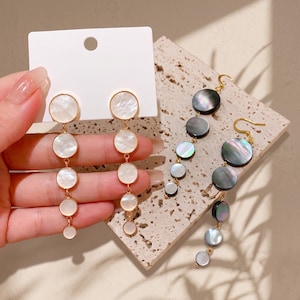 May include: Two pairs of drop earrings featuring a vertical arrangement of five circular discs in varying sizes. One pair consists of iridescent white shell discs with gold-tone metal connectors, while the other pair features dark grey shell discs.