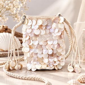 Peut inclure: Un petit sac seau recouvert de disques de coquillages circulaires iridescents et superposés dans des tons crème et rose pâle. Le sac présente une base en tissu beige et une bandoulière en corde tressée avec des disques assortis aux extrémités.