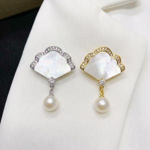 Op de afbeelding: Twee waaiervormige broches, één in zilverkleurig en één in goudkleurig metaal. Elke broche heeft een centrale inleg van iriserend parelmoer, een rand van kleine heldere steentjes en een ronde witte parel die aan een helder steentje hangt.
