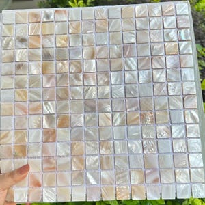 Puede incluir: Una lámina de mosaico cuadrado compuesta por numerosas baldosas cuadradas pequeñas. Las baldosas exhiben un brillo nacarado con variaciones de tonos blancos, crema y marrón claro. El mosaico es adecuado para diversas aplicaciones decorativas.