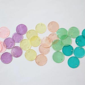 Puede incluir: Un conjunto de cuentas redondas translúcidas de colores pastel: rosa, morado, amarillo, melocotón, verde y turquesa. Cada cuenta tiene un pequeño agujero cerca del borde. Las cuentas están dispersas sobre una superficie blanca.