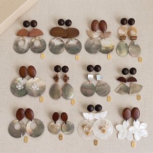 May include: A collection of twelve pairs of drop earrings featuring dark brown wooden studs and various shapes of iridescent shell pendants. The designs include circular, teardrop, and floral motifs, some accented with small white shell flowers or starfish.