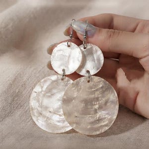 May include: A pair of drop earrings featuring two stacked circular discs made of iridescent white shell. The earrings are attached to silver-toned metal fishhook wires, with a smaller disc positioned above a larger disc on each earring.