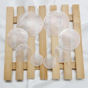 Può includere: Una collezione di pezzi rotondi e iridescenti di conchiglia di madreperla con piccoli fori, disposti su un reticolo di legno marrone chiaro. I gusci variano di dimensioni, con una lucentezza perlacea, ideali per la creazione di gioielli o progetti artigianali.