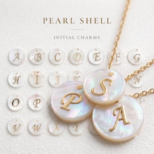 Op de afbeelding: Een collectie ronde, iriserende parelmoer bedels met goudkleurige gegraveerde letters. De tekst PEARL SHELL INITIAL CHARMS staat boven de bedels. Twee bedels zijn bevestigd aan goudkleurige metalen kettingen.