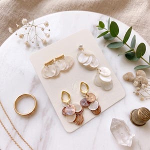 May include: Three pairs of earrings displayed on a rectangular card. One pair features white circular shell discs with gold accents, another has white shell discs with a pearl stud, and the third pair has gold teardrop tops with brown and white shell discs.