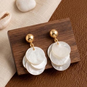 May include: A pair of gold-tone stud earrings featuring a polished spherical top and three dangling, circular, iridescent white shell discs. The earrings are displayed on a rectangular piece of dark brown wood with a visible grain pattern.