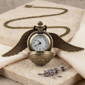 Puede incluir: Un reloj de bolsillo de color bronce con una cadena, con una esfera redonda y alas decorativas. El reloj tiene una esfera blanca con números y agujas negras. La palabra "EYNAR" está en la esquina superior derecha.