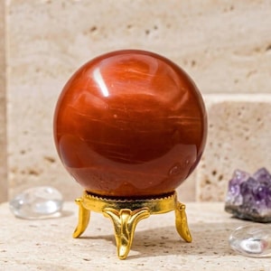 May include: A polished, spherical red stone orb with subtle swirling patterns, resting on a small, three-legged gold-toned metal stand. The smooth, glossy surface of the sphere reflects light, highlighting its deep reddish-brown color and natural texture.