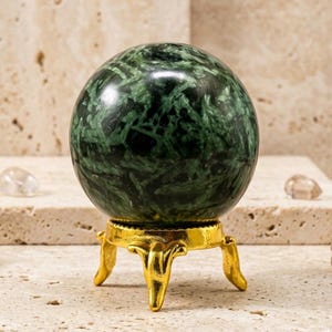 Puede incluir: Una esfera de color verde oscuro pulido con intrincados patrones cristalinos negros y verde claro, que descansa sobre un pequeño soporte de metal dorado de tres patas. La esfera presenta una superficie lisa y reflectante con inclusiones minerales.