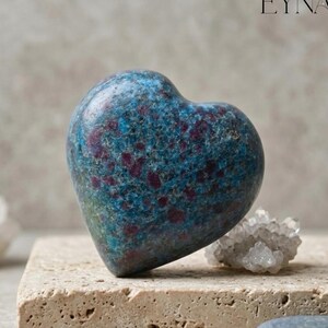 May include: A heart-shaped stone featuring a mottled texture with deep blue and vibrant magenta-red inclusions. The polished surface displays a mix of dark and light blue tones speckled with distinct reddish-purple spots throughout the piece.