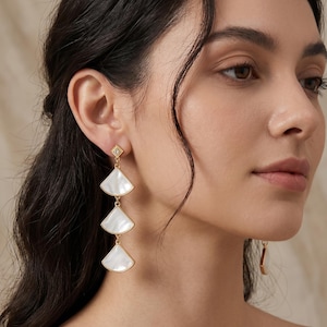 Puede incluir: Pendientes dorados con una parte superior en forma de diamante y tres colgantes blancos en forma de abanico. Los pendientes son colgantes y tienen un acabado pulido. El diseño es elegante y adecuado para ocasiones formales.