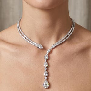 May include: A diamond necklace with a double-strand design that wraps around the neck. The necklace has a snake-like head and a series of pear-shaped diamonds that hang down the chest. The necklace is made of white metal and is set with many small, sparkling diamonds.