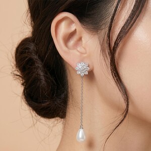 May include: A silver earring featuring a flower-shaped stud set with small, clear stones. A fine chain supports a pear-shaped, white pearl. The earring is worn on an ear, with the person's hair styled up.