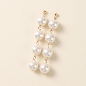 May include: A pair of dangling earrings featuring six large, round, white faux pearls suspended from gold-colored posts and chains. The earrings have a classic, elegant design, suitable for formal or everyday wear.