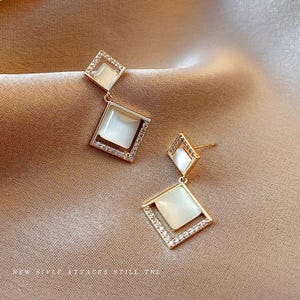 May include: Gold-toned, geometric drop earrings. Each earring features two stacked, diamond-shaped frames. The inner frame holds a square, opalescent stone. The outer frame is encrusted with small, clear crystals. The earrings dangle from a post. The background is a tan fabric.