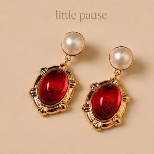 Könnte beinhalten: Ein Paar goldfarbene Ohrringe mit einem Perlenstecker und einem roten, oval geformten Edelstein. Die Edelsteine sind in einem dekorativen Goldrahmen gefasst. Der Text "little pause" befindet sich oben.