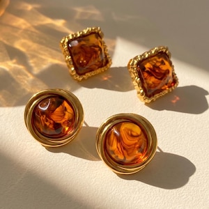 May include: Four vintage-style earrings. Two pairs of earrings are visible. One pair is square with a gold-colored, textured frame and a marbled amber-colored center. The other pair is round with a gold-colored frame and a marbled amber center.