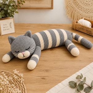 Puede incluir: Un juguete de gato tejido a ganchillo con un patrón de rayas grises y crema. El gato tiene una cabeza redondeada con orejas puntiagudas, ojos cerrados, nariz rosa y bigotes blancos. Su cuerpo es alargado con extremidades a rayas y cola.