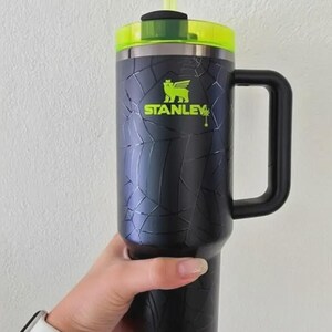 Pode incluir: Caneca Stanley preta e prateada com al&ccedil;a, tampa verde e canudo verde. A caneca tem um design de padr&atilde;o rachado e o logotipo Stanley em verde. A caneca &eacute; segurada por uma pessoa.