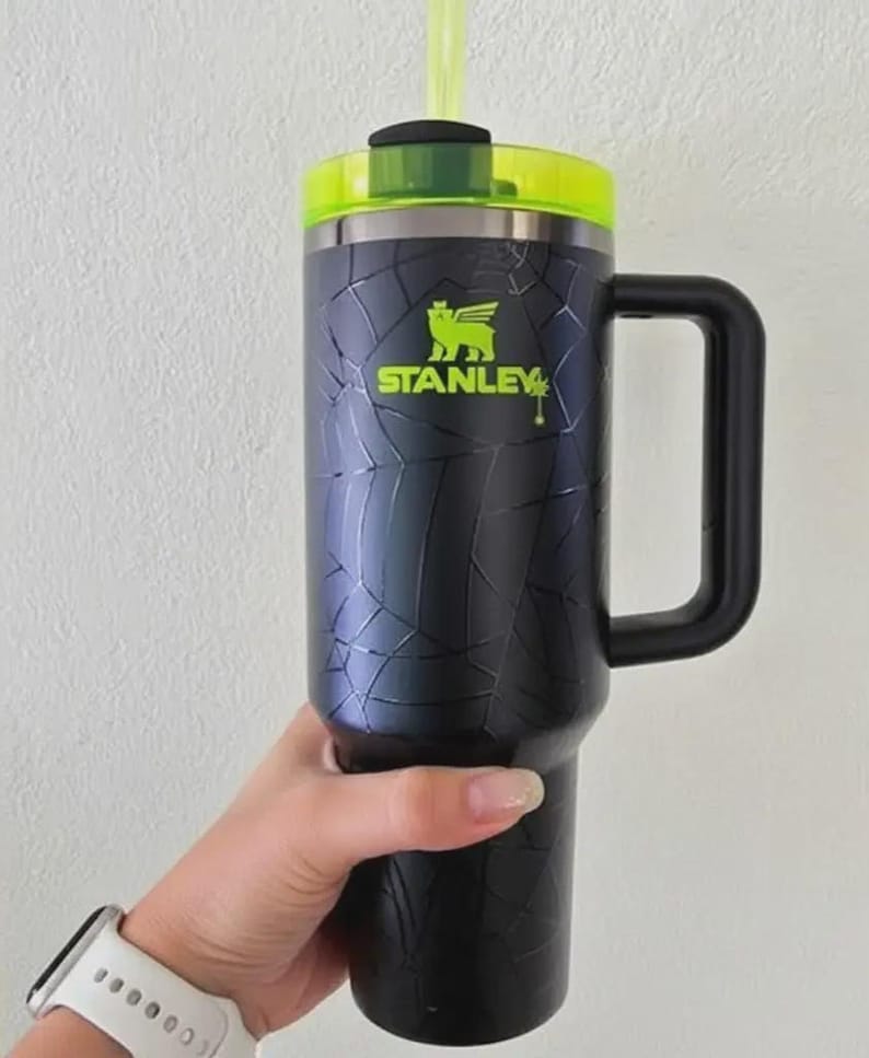 Pode incluir: Caneca Stanley preta e prateada com al&ccedil;a, tampa verde e canudo verde. A caneca tem um design de padr&atilde;o rachado e o logotipo Stanley em verde. A caneca &eacute; segurada por uma pessoa.