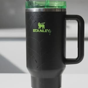 Pode incluir: Caneca Stanley Quencher preta com tampa e canudo verdes. A caneca tem uma al&ccedil;a e um design de teia de aranha. O logotipo da Stanley &eacute; verde. Uma etiqueta branca est&aacute; na parte inferior da caneca.