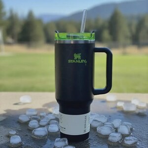 Pode incluir: Um copo t&eacute;rmico Stanley preto com tampa e al&ccedil;a verdes, com o logotipo Stanley em verde. Um canudo transparente se estende da tampa. O copo est&aacute; sobre uma superf&iacute;cie molhada cercada por cubos de gelo. O fundo mostra um campo verde e &aacute;rvores.