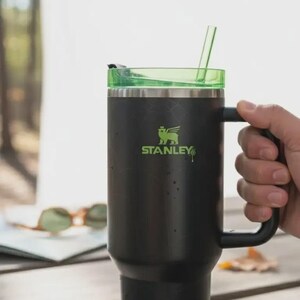 Pode incluir: Caneca Stanley preta com tampa e canudo verdes. A caneca tem uma al&ccedil;a e o logotipo Stanley em verde. A caneca est&aacute; em uma mesa de madeira ao ar livre.
