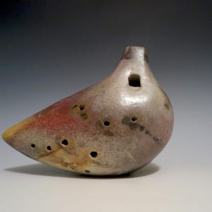 May include: A ceramic ocarina in a bird-like shape. The instrument is primarily gray with red and yellow accents. It has several finger holes and a square opening near the top. The ocarina sits on a white surface.