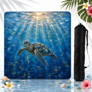May include: A square beach towel featuring an impressionistic painting of a sea turtle swimming in deep blue water with light rays filtering from above. Beside the towel is a black cylindrical carrying bag with a shoulder strap.