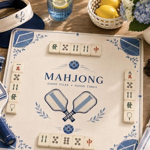 Puede incluir: Un tablero de juego cuadrado con fondo crema, detalles florales azules y el texto "MAHJONG GOOD TILES GOOD TIMES" en el centro. El tablero presenta ilustraciones de dos palas de pickleball y est&aacute; rodeado por filas de fichas de mahjong.