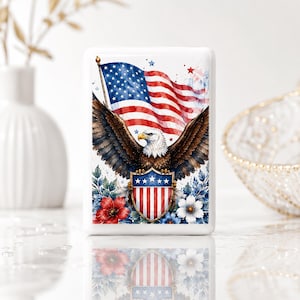 May include: A rectangular white magnet featuring a watercolor illustration of a bald eagle with wings spread wide. The eagle is positioned in front of a waving American flag and holds a shield with red and white stripes and blue stars.