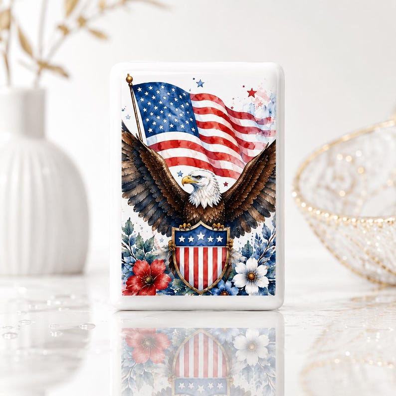 May include: A rectangular white magnet featuring a watercolor illustration of a bald eagle with wings spread wide. The eagle is positioned in front of a waving American flag and holds a shield with red and white stripes and blue stars.