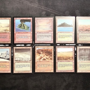 May include: A collection of ten rectangular trading cards arranged in two rows of five. Each card features a unique landscape illustration, a light-colored border, and printed text detailing game mechanics, including mana pool information.