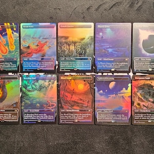 Könnte beinhalten: Eine Sammlung von zehn Magic: The Gathering Sammelkarten, angeordnet in zwei Reihen. Jede Karte zeigt vollflächige Landschaftsillustrationen mit lebendigen, holografischen Folienoberflächen und Textfeldern zur Spielmechanik.