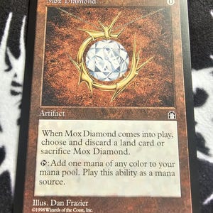 Könnte beinhalten: Magic: The Gathering Karte "Mox Diamond" mit einem Diamant-Anhänger in Goldfassung auf braunem Hintergrund. Der Text beschreibt seine Funktion als Artefakt im Spiel.