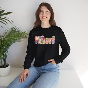 Può includere: Felpa nera con la scritta "Mimi" in un colorato design patchwork. La parola è composta da vari motivi e colori, tra cui rosso, blu, giallo e verde. La persona indossa jeans blu.