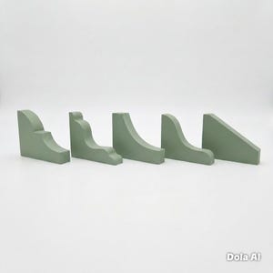 May include: Five sage green wooden corner brackets arranged in a row. Each bracket features a distinct profile, including curved, stepped, and angled designs. The pieces have a smooth, matte finish and are positioned upright on a flat surface.