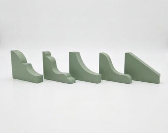 May include: Five sage green wooden corner brackets arranged in a row. Each bracket features a distinct profile, including curved, stepped, and angled designs. The pieces have a smooth, matte finish and are positioned upright on a plain surface.