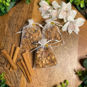 May include: Three small, sheer bags filled with dried fruit and spices, tied with white ribbon, sit on a wooden surface. Several cinnamon sticks are arranged nearby. White flowers with pink stripes add a decorative touch.