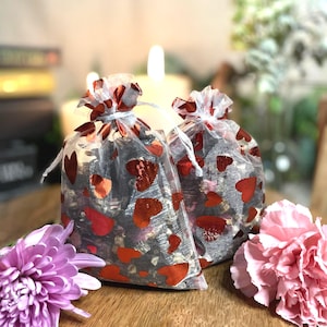 May include: Two clear mesh bags filled with dark contents and red heart-shaped confetti. The bags are tied with white string and sit on a wooden surface. Pink and purple flowers are in the foreground, with lit candles in the background.