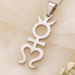 May include: A silver-colored pendant necklace featuring a stylized astrological symbol. The pendant hangs from a delicate chain and is displayed on a cream-colored fabric. A small white rose and sprigs of white flowers are in the lower right corner.