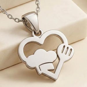 May include: A silver-colored pendant necklace featuring a heart-shaped design with a chef's hat and fork. The pendant hangs from a delicate chain. The necklace is displayed on a cream-colored surface.