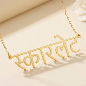 May include: A gold-colored necklace with a chain and a pendant. The pendant features the word "Scarlett" written in a stylized script. The necklace is displayed on a light-colored surface, with a white flower in the background.