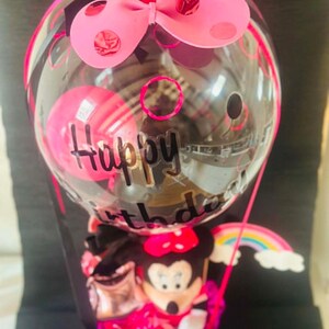 May include: A clear balloon with black Happy Birthday text, topped with a pink bow with red polka dots. Inside the balloon is a smaller pink balloon. Below, a cartoon mouse figure sits in a base with a small rainbow cutout attached to the side.