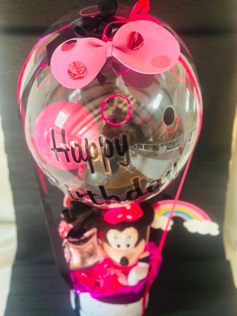 May include: A clear balloon with black Happy Birthday text, topped with a pink bow with red polka dots. Inside the balloon is a smaller pink balloon. Below, a cartoon mouse figure sits in a base with a small rainbow cutout attached to the side.