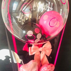 May include: A decorative gift arrangement featuring a large clear balloon with pink and black polka dots, containing smaller pink and black balloons. Below, a white cylindrical base holds two small heart-shaped pink balloons and a large peach bow.