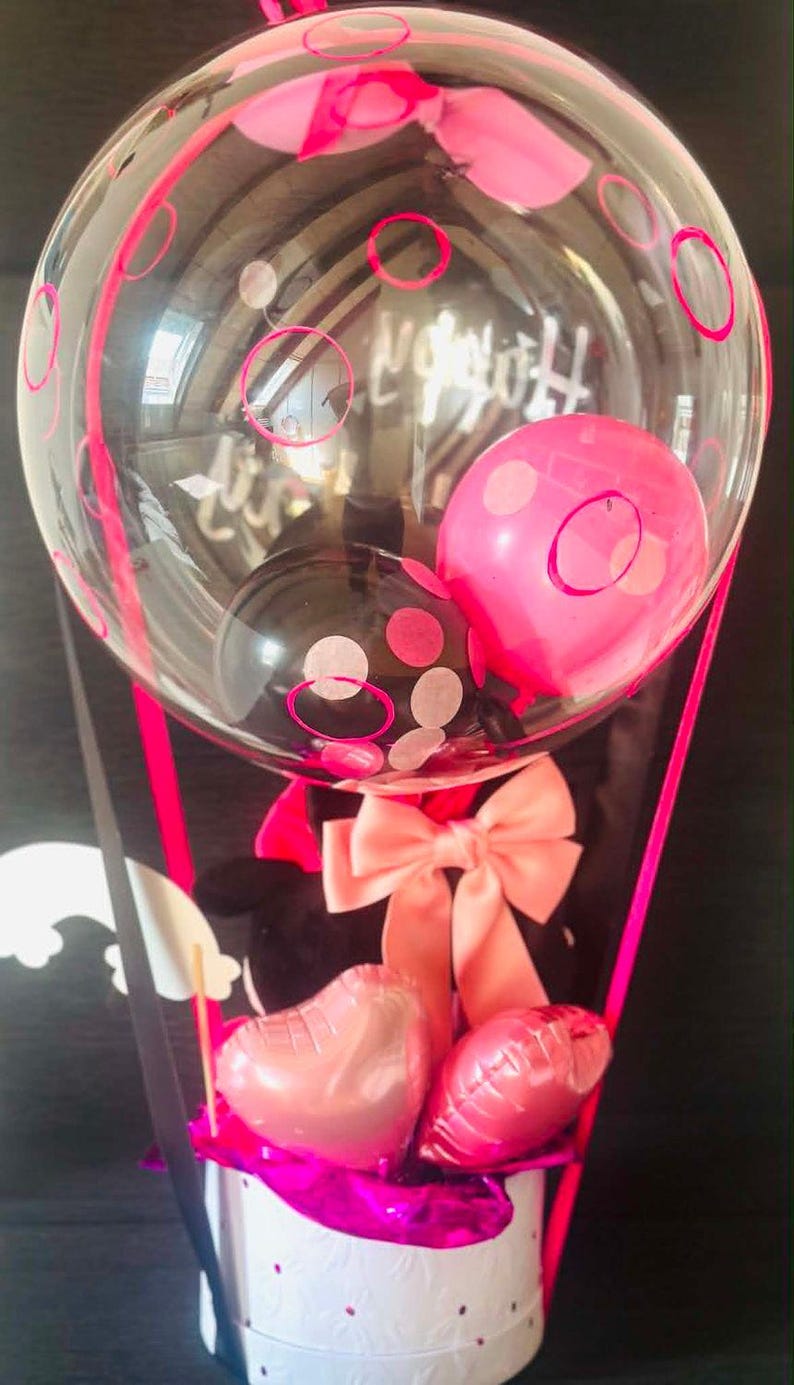 May include: A decorative gift arrangement featuring a large clear balloon with pink and black polka dots, containing smaller pink and black balloons. Below, a white cylindrical base holds two small heart-shaped pink balloons and a large peach bow.