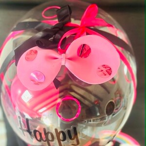 May include: A clear, round balloon decorated with a large pink bow featuring metallic pink polka dots, black and neon pink ribbons, and the word Happy printed in black script. Inside the balloon, smaller pink balloons are visible.