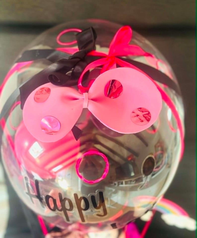 May include: A clear, round balloon decorated with a large pink bow featuring metallic pink polka dots, black and neon pink ribbons, and the word Happy printed in black script. Inside the balloon, smaller pink balloons are visible.